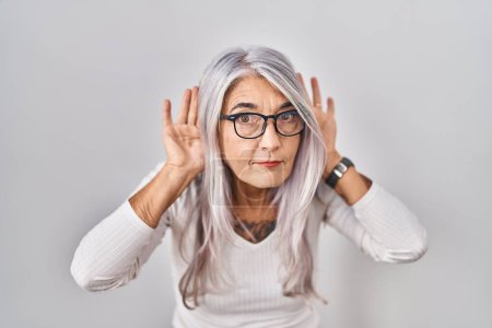 Foto de Mujer de mediana edad con el pelo gris de pie sobre fondo blanco tratando de escuchar ambas manos en el gesto de la oreja, curioso por chismes. problema auditivo, sordo - Imagen libre de derechos