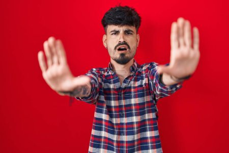 Foto de Joven hombre hispano con barba de pie sobre fondo rojo haciendo gesto de stop con palmas de las manos, expresión de enojo y frustración - Imagen libre de derechos