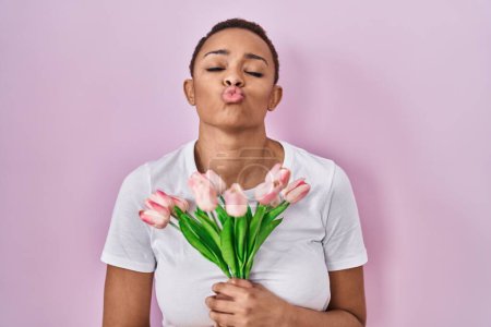 Téléchargez les photos : Belle femme afro-américaine tenant bouquet de fleurs de tulipes roses regardant la caméra souffler un baiser étant belle et sexy. expression d'amour. - en image libre de droit