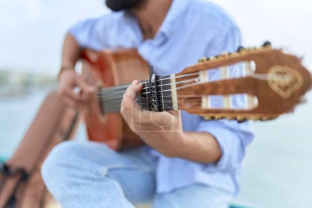 Photo for Young hispanic man musician playing classical guitar sitting on balustrade at seaside - Royalty Free Image