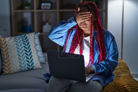 Téléchargez les photos : Femme afro-américaine aux cheveux tressés utilisant ordinateur portable la nuit souriant et riant avec la main sur le visage couvrant les yeux pour la surprise. concept aveugle. - en image libre de droit