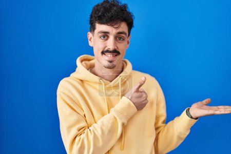 Photo for Hispanic man standing over blue background showing palm hand and doing ok gesture with thumbs up, smiling happy and cheerful - Royalty Free Image