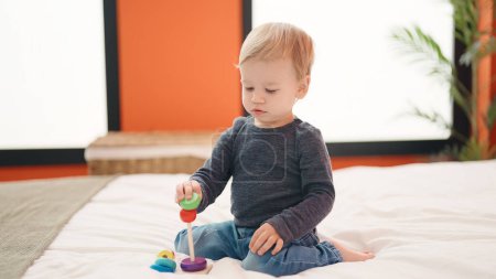 Foto de Adorable niño rubio jugando con aros de juguete sentado en la cama en el dormitorio - Imagen libre de derechos