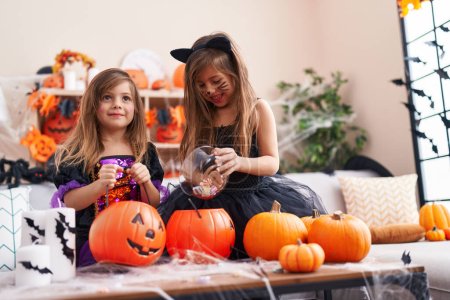 Foto de Adorables chicas teniendo fiesta de Halloween poniendo caramelos en la cesta de calabaza en casa - Imagen libre de derechos