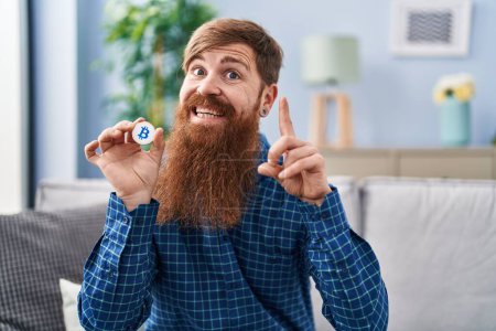 Foto de Hombre caucásico con barba larga sosteniendo bitcoin moneda virtual sorprendido con una idea o pregunta señalando el dedo con la cara feliz, número uno - Imagen libre de derechos