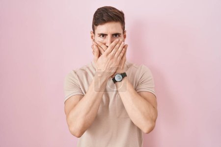 Foto de Hombre hispano con barba de pie sobre fondo rosa impactó cubriendo la boca con las manos por error. concepto secreto. - Imagen libre de derechos