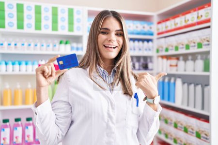 Téléchargez les photos : Jeune femme hispanique travaillant à la pharmacie pharmacie tenant carte de crédit pointant le pouce vers le côté souriant heureux avec la bouche ouverte - en image libre de droit