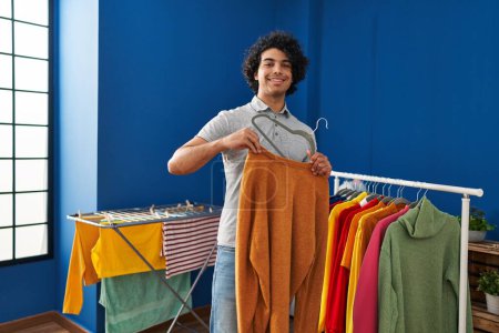 Téléchargez les photos : Young hispanic man smiling confident hanging clothes on rack at laundry room - en image libre de droit