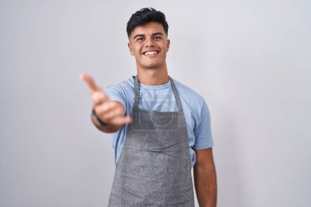 Photo for Hispanic young man wearing apron over white background smiling cheerful offering palm hand giving assistance and acceptance. - Royalty Free Image