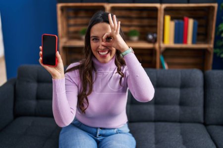 Téléchargez les photos : Young brunette woman holding smartphone showing blank screen smiling happy doing ok sign with hand on eye looking through fingers - en image libre de droit