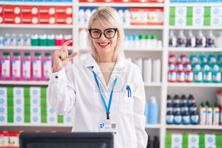 Foto de Young caucasian woman working at pharmacy drugstore smiling and confident gesturing with hand doing small size sign with fingers looking and the camera. measure concept. - Imagen libre de derechos