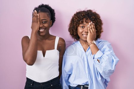 Foto de Dos mujeres africanas de pie sobre fondo rosa cubriendo un ojo con la mano, sonrisa confiada en la cara y emoción sorpresa. - Imagen libre de derechos