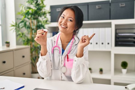 Foto de Joven asiática mujer vistiendo médico uniforme y estetoscopio sonriendo feliz señalando con la mano y el dedo a un lado - Imagen libre de derechos