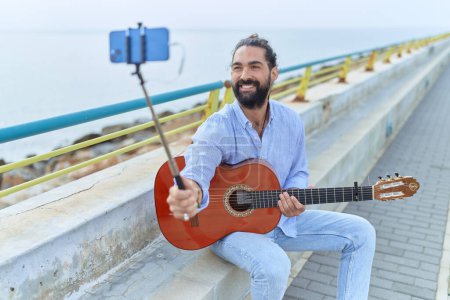 Photo for Young hispanic man musician holding classical guitar having video call at seaside - Royalty Free Image