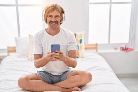 Foto de Young blond man listening to music sitting on bed at bedroom - Imagen libre de derechos