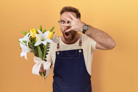 Foto de Hombre de mediana edad con barba floristería sosteniendo flores asomándose en shock cubriendo la cara y los ojos con la mano, mirando a través de los dedos con expresión avergonzada. - Imagen libre de derechos