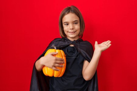 Foto de Pequeño niño hispano usando una capa y sosteniendo la calabaza de Halloween sonriendo feliz señalando con la mano y el dedo hacia un lado - Imagen libre de derechos