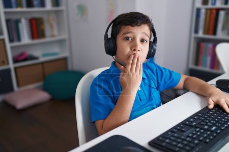 Foto de Pequeño niño hispano usando computadora portátil en la escuela cubriendo la boca con la mano, sorprendido y temeroso de equivocarse. expresión sorprendida - Imagen libre de derechos