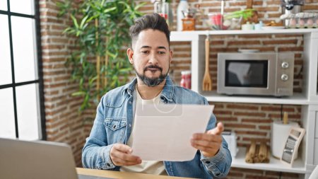 Photo for Young latin man using laptop reading document at dinning room - Royalty Free Image
