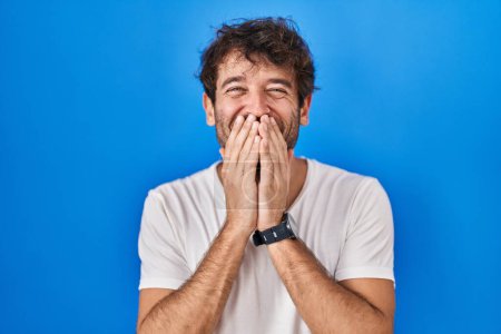 Téléchargez les photos : Jeune homme hispanique debout sur fond bleu rire et rire embarrassé couvrant la bouche avec les mains, commérages et concept scandale - en image libre de droit