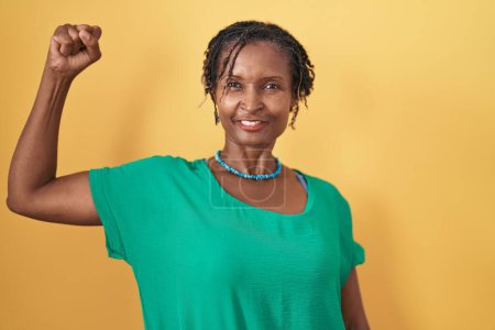 Foto de Mujer africana con rastas de pie sobre fondo amarillo persona fuerte que muestra el músculo del brazo, confiado y orgulloso de poder - Imagen libre de derechos