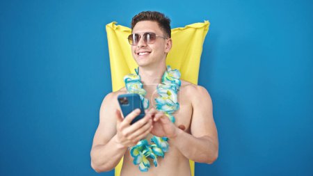 Téléchargez les photos : Jeune homme hispanique touriste utilisant le smartphone détendu sur hamac gonflable de piscine sur fond bleu isolé - en image libre de droit