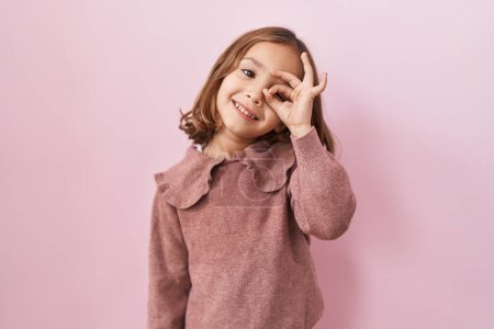 Foto de Pequeña chica hispana de pie sobre fondo rosa sonriendo feliz haciendo signo ok con la mano en el ojo mirando a través de los dedos - Imagen libre de derechos