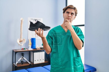 Photo for Young man working at physiotherapy clinic holding shoe serious face thinking about question with hand on chin, thoughtful about confusing idea - Royalty Free Image