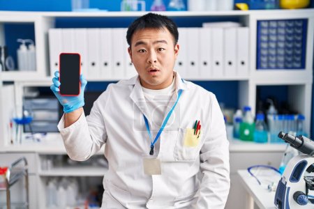 Photo for Young chinese man working at scientist laboratory holding smartphone scared and amazed with open mouth for surprise, disbelief face - Royalty Free Image