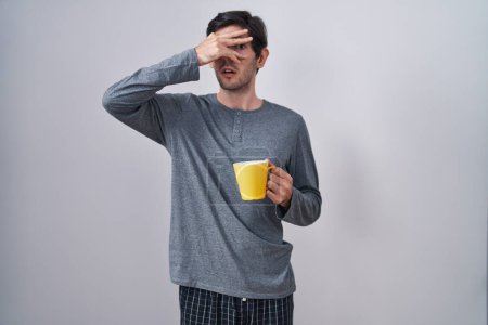Téléchargez les photos : Jeune homme hispanique portant un pyjama boire une tasse de café en regardant dans le choc couvrant le visage et les yeux avec la main, regardant à travers les doigts avec une expression embarrassée. - en image libre de droit
