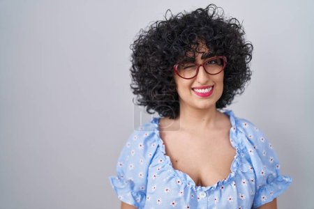 Photo for Young brunette woman with curly hair wearing glasses over isolated background winking looking at the camera with sexy expression, cheerful and happy face. - Royalty Free Image