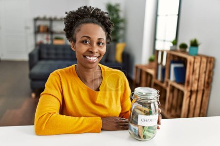 Foto de Mujer afroamericana sentada en mesa sosteniendo frasco de caridad con billetes de rand en casa - Imagen libre de derechos