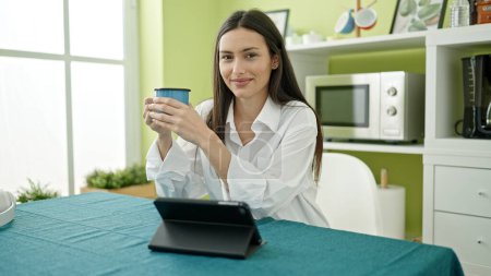 Foto de Joven hermosa mujer hispana usando touchpad bebiendo café sentado en la mesa en el comedor - Imagen libre de derechos