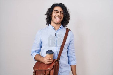 Foto de Hombre hispano con el pelo rizado bebiendo una taza de café para llevar sacando la lengua feliz con expresión divertida. concepto de emoción. - Imagen libre de derechos