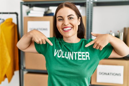 Photo for Young beautiful hispanic woman smiling confident pointing with fingers to volunteer t shirt at charity center - Royalty Free Image