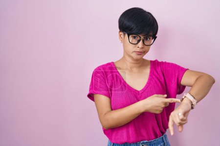 Téléchargez les photos : Jeune femme asiatique avec les cheveux courts debout sur fond rose à la hâte pointant pour regarder le temps, l'impatience, bouleversé et en colère pour délai - en image libre de droit