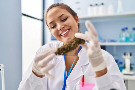 Foto de Joven hermosa mujer hispana científica sonriendo confiada sosteniendo marihuana en el laboratorio - Imagen libre de derechos
