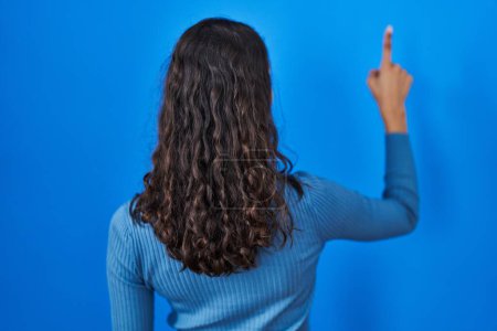 Téléchargez les photos : Jeune femme brésilienne debout sur fond bleu isolé posant vers l'arrière pointant vers l'avant avec la main du doigt - en image libre de droit