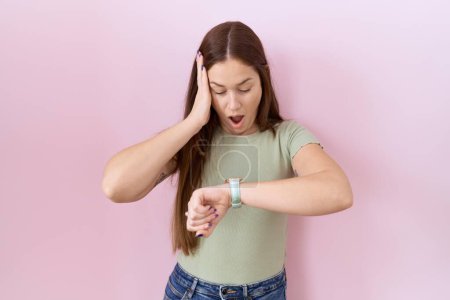 Téléchargez les photos : Belle femme brune debout sur fond rose regardant le temps de la montre inquiet, peur de se faire tard - en image libre de droit