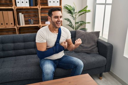 Photo for Young hispanic man with beard wearing arm on sling sitting at therapy consult screaming proud, celebrating victory and success very excited with raised arms - Royalty Free Image