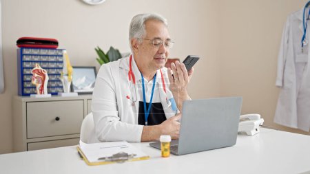 Foto de Hombre de mediana edad con médico de pelo gris que envía un mensaje de voz con teléfono inteligente en la clínica - Imagen libre de derechos