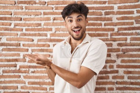 Téléchargez les photos : Homme arabe avec barbe debout sur le mur de briques arrière-plan pointant de côté avec les mains ouvertes paumes montrant l'espace de copie, présentant la publicité souriant heureux heureux - en image libre de droit