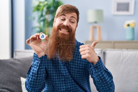 Photo for Caucasian man with long beard holding virtual currency bitcoin pointing thumb up to the side smiling happy with open mouth - Royalty Free Image