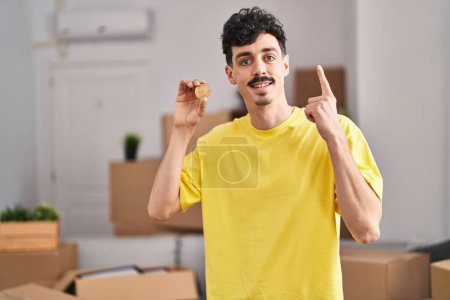 Téléchargez les photos : Homme hispanique tenant Bitcoin à la nouvelle maison souriant et riant fort à haute voix parce que drôle blague folle. - en image libre de droit