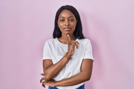 Foto de Mujer joven africana con camiseta blanca casual pensando concentrada en la duda con el dedo en la barbilla y mirando hacia arriba preguntándose - Imagen libre de derechos