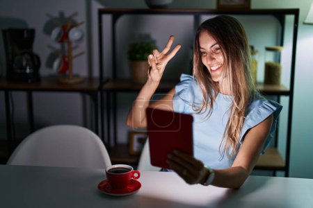 Téléchargez les photos : Jeune femme hispanique utilisant pavé tactile assis sur la table la nuit souriant en regardant la caméra montrant les doigts faisant signe de victoire. numéro deux. - en image libre de droit