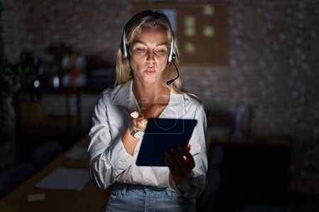 Foto de Young blonde woman working at the office at night looking at the camera blowing a kiss with hand on air being lovely and sexy. love expression. - Imagen libre de derechos