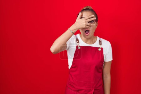 Foto de Mujer hispana joven vistiendo delantal de camarera sobre fondo rojo asomándose en shock cubriendo la cara y los ojos con la mano, mirando a través de los dedos con expresión avergonzada. - Imagen libre de derechos