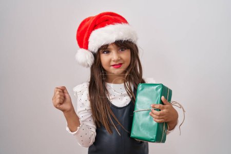 Téléchargez les photos : Petite fille hispanique portant un chapeau de Noël et tenant des cadeaux criant fier, célébrant la victoire et le succès très excité avec le bras levé - en image libre de droit