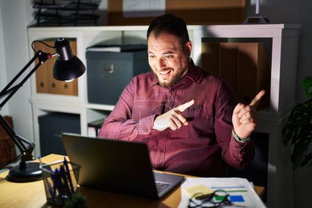 Photo for Plus size hispanic man with beard working at the office at night smiling and looking at the camera pointing with two hands and fingers to the side. - Royalty Free Image